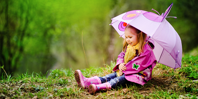 Насмешена девојчица са кишобраном у кишном капуту седи на отвореном