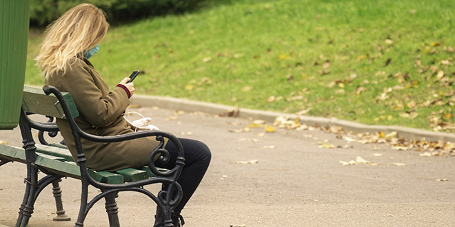 Жена са телефоном у рукама седи на клупи у парку